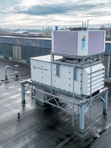 Luftaufnahme eines großen, auf einem Fabrikdach installierten Lüftungssystems. Die Anlage steht auf einer Metallstruktur und ist vorwiegend in Weiß gehalten, mit einem oberen Gitter in Silber. Das Dach ist nass, möglicherweise nach einem Regenschauer. Im Hintergrund ist eine städtische Umgebung mit entfernten Gebäuden und leicht bewölktem Himmel zu erkennen. | © INFRANORM®