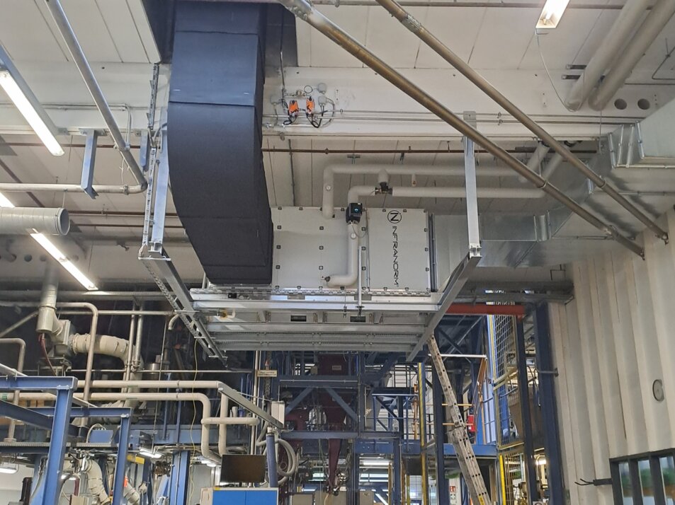 Konditionierte Hallenluft für angenehmes Arbeitsklima. | © Infranorm, Wels (AT) Industrielle Produktionshalle mit komplexen Rohrleitungen und einer großen, schwarzen Lüftungsanlage an der Decke. Unten arbeitet eine Person an einem Tisch mit einem Laptop, umgeben von Maschinen und technischen Geräten. Im Hintergrund sind Leitern und weitere technische Strukturen zu sehen. | © Infranorm, Wels (AT)