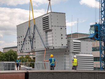Einheben eines Kubus des RLT-Geräts von bösch per Kran auf das Hallendach; Monteure sichern das Bauteil. | © Walter Bösch GmbH & Co KG