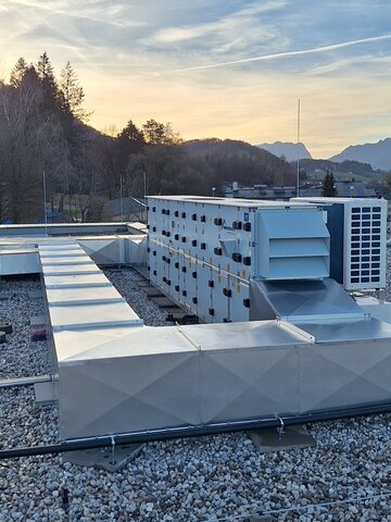 Ein Dach mit mehreren Klimaanlagen-Einheiten in verschiedenen Größen und Formen bei Sonnenuntergang. Die Anlagen sind auf einem mit Kies bedeckten Dach platziert, das sich vor einem Hintergrund mit bewaldeten Bergen erstreckt. Der Himmel ist leicht bewölkt und färbt sich orange durch die untergehende Sonne.